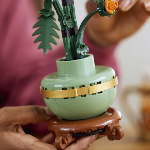 Close-up do vaso verde sage do LEGO 10368 com aro dourado e base de madeira sendo segurado pela mão