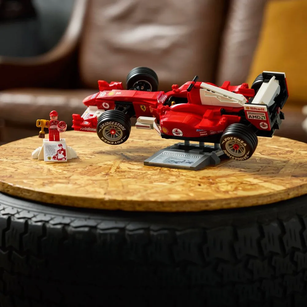 LEGO Icons 11375 Ferrari F2004 montado sobre mesa de madeira ao lado da minifigura do Schumacher com troféu, cenário com pneu de F1 e poltrona de couro